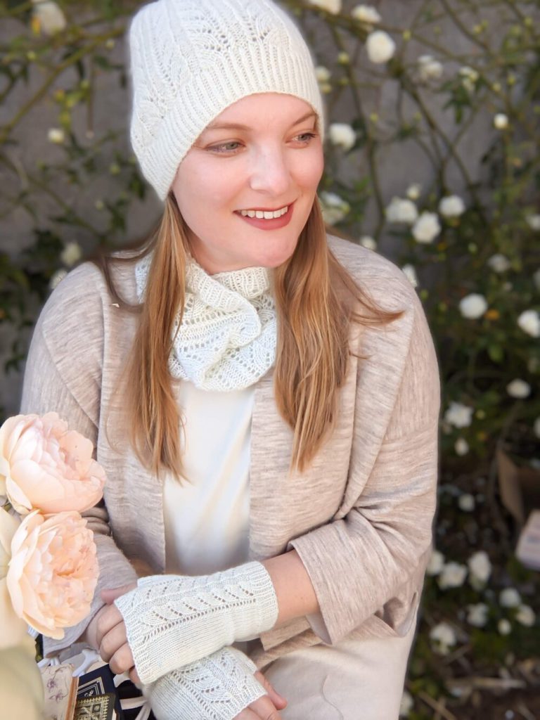 A blonde woman with long hair looks off to the side while wearing a tan sweater and lacy knit hat, cowl, and mitts. The hat, cowl, and mitts are all in a very pale mint color.
