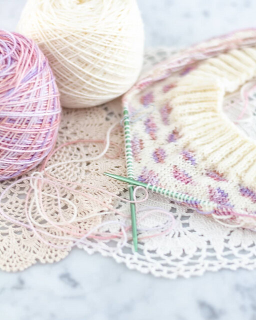 A stranded colorwork cowl in white and variegated pink-purple is laid flat on a white marble countertop. Test knitting helps me ensure these knit projects turn out smoothly.