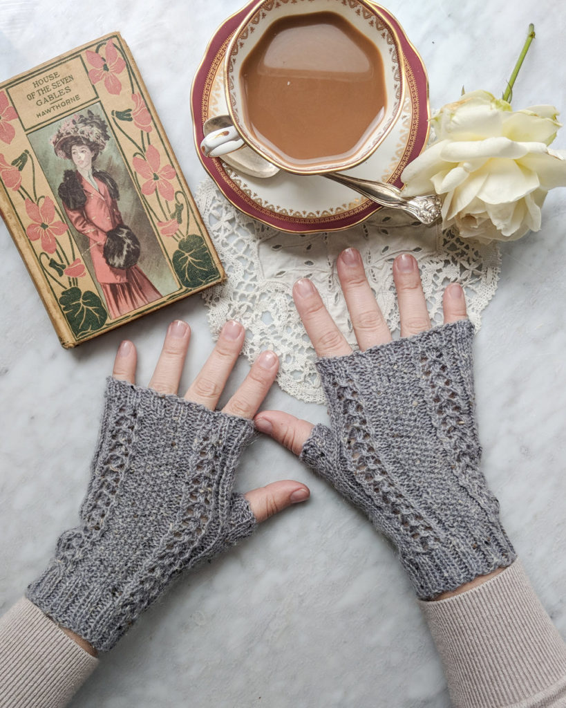 A pair of small hands wearing gray, fingerless mitts are splayed out on a white marble countertop with an antique book, a teacup, and a white rose.