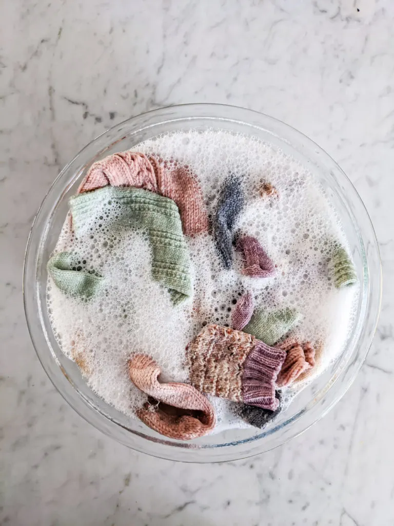 A bowl full of pastel socks and soap suds on a white marble countertop. These hand knit socks require a little care, but this guide lays it all out for you.