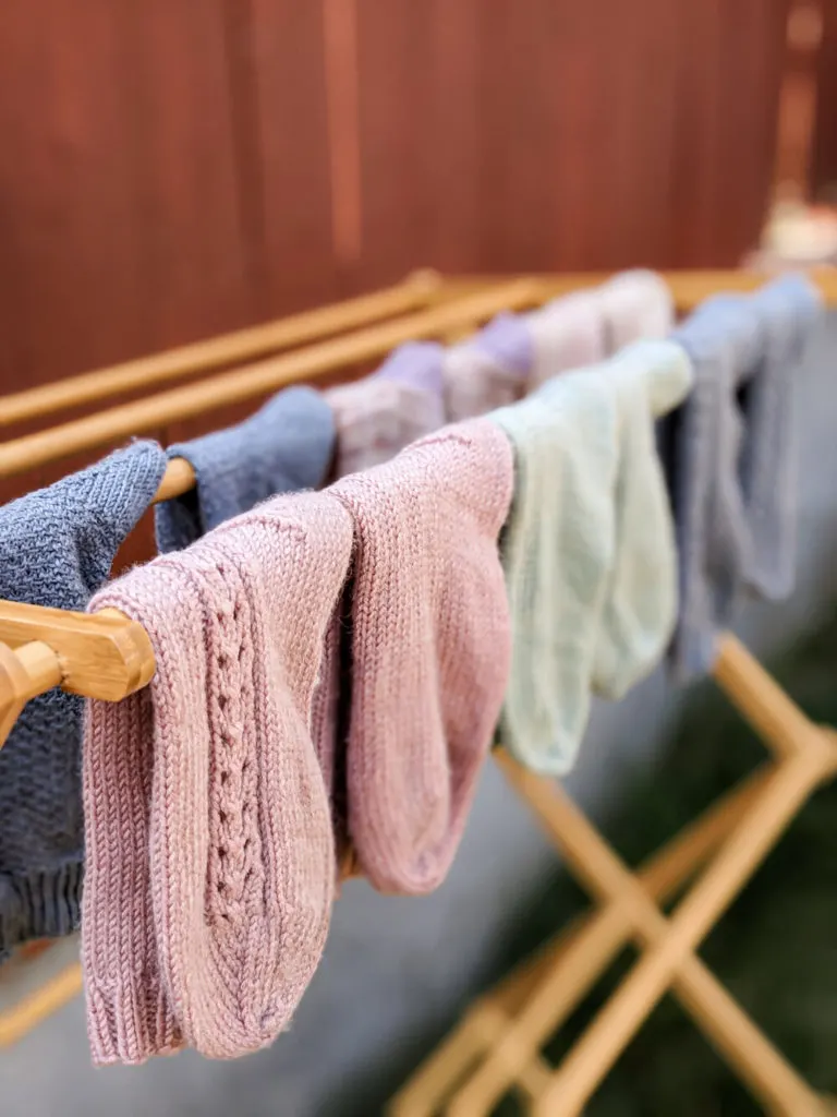 Several pairs of hand knit socks in pastel colors dry on a bamboo rack outside.