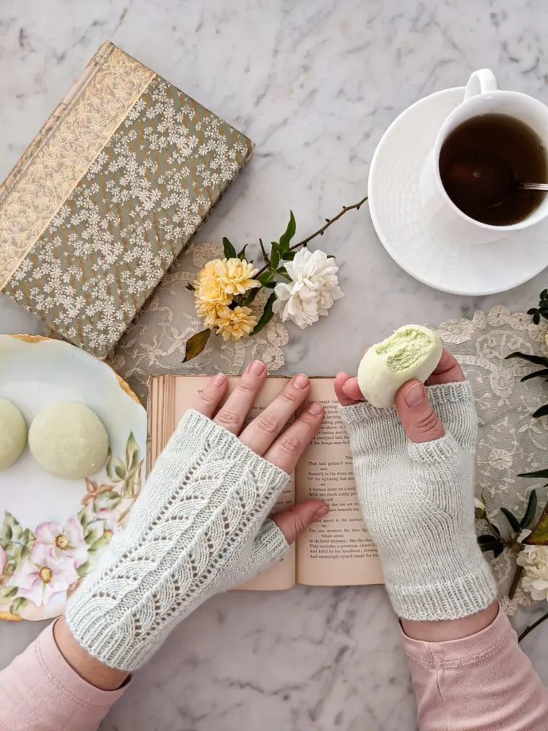 A pair of small, plump hands wears very pale mint-green mitts with an arrow-shaped lace pattern up the back. The right hand holds a green tea mochi ice cream ball. Two more balls sit on a floral gilded plate, along with some antique books, tiny roses, and a white teacup full of tea.