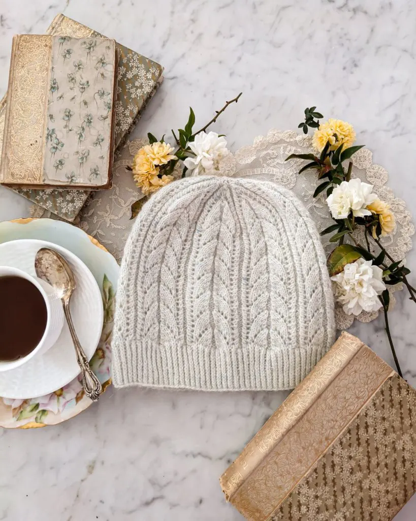 A very pale mint green hat with an arrow-shaped lace pattern sits flat on a white marble countertop, along with some antique books, tiny roses, and a white teacup full of tea.