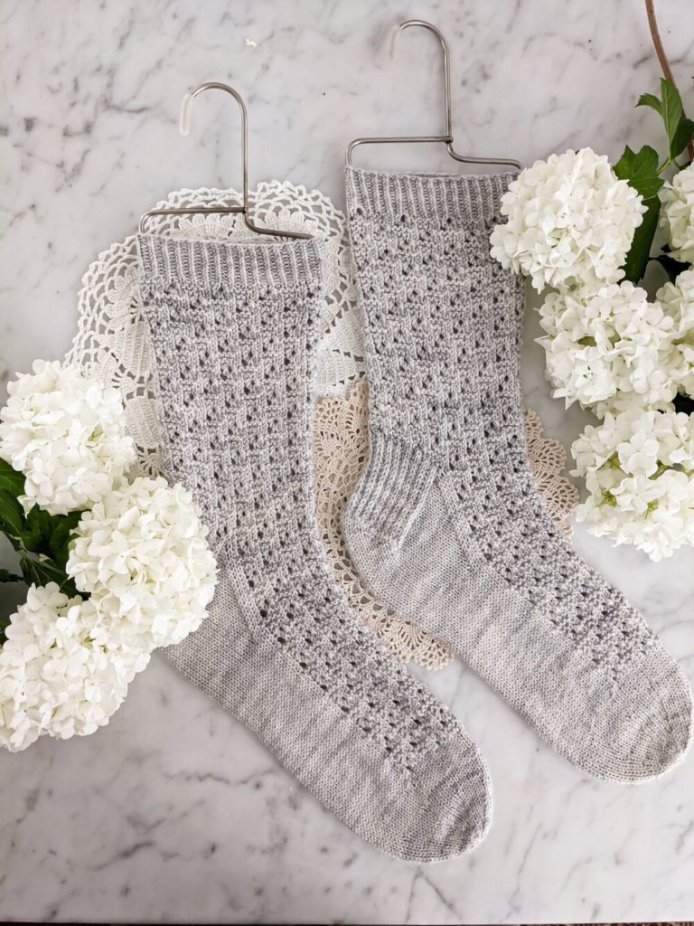 A pair of light blue-gray, textured socks is laid flat on a white marble countertop with some white hydrangea next to them.