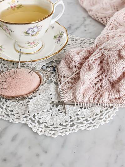 A pink, lacy sweater sleeve on steel needles is laid out on a white marble countertop with an antique pincushion and a white teacup filled with chamomile tea. Test knitting helps me figure out whether details, like lace patterns in my designs, line up in a way that makes sense for knitters.