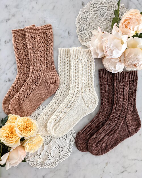 Three socks in pink, white, and brown are laid out on a white marble countertop with clusters of peach and yellow roses. These socks represent a triple test knit opportunity.