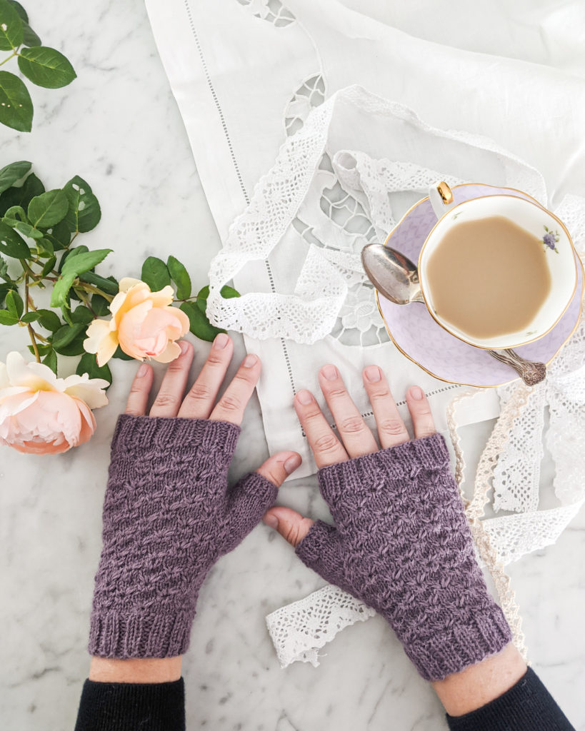 A pair of purple, fingerless mitts with an all-over textured design on two hands splayed out across a white marble countertop. There are pale pink roses to the left, a white lacy tablecloth underneath the mitts/hands, and a lavender teacup filled with milky tea on the right.