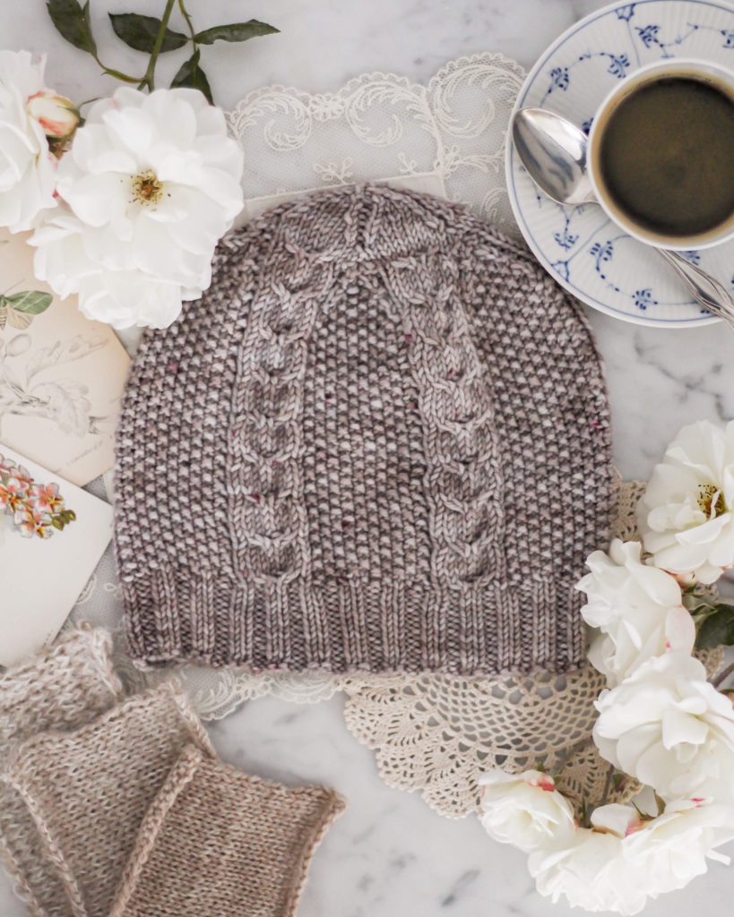 A gray knit hat with cabled columns and seed stitch sits flat on a white marble countertop, surrounded by a teacup full of coffee, white roses, swatches of tan knit fabric, and antique paper ephemera.