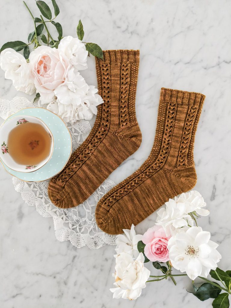 The Creme Brulee Socks, a pair of caramel-colored socks with eyeled and seed stitch panels, are laid flat on a white marble surface with both toes pointed left. They're surrounded by pink and white roses and a mint-colored teacup.