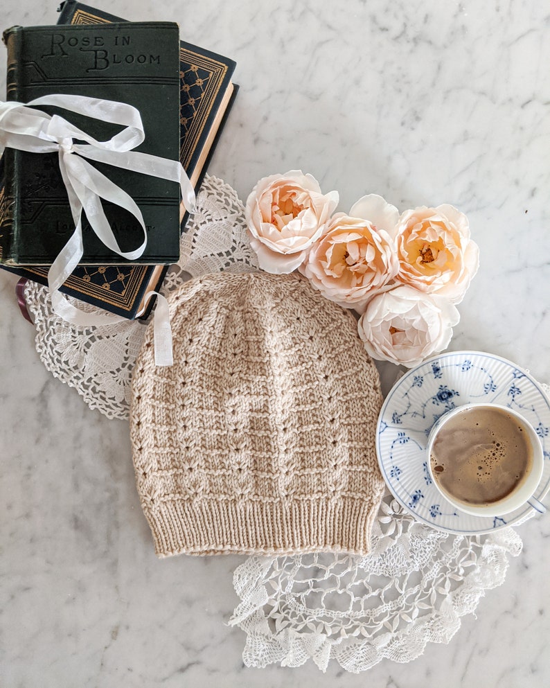 The Gingersnap Hat, a cream-colored hat with lots of texture, sits flat on a white marble countertop surrounded by green antique books, peach roses, and a blue and white tea cup.