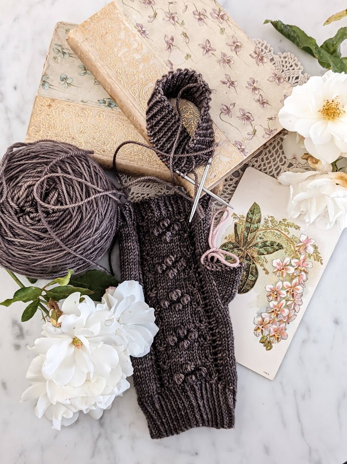 A mostly-finished fingerless mitt and the cuff for the second mitt sit surrounded by antique books with floral and gilded covers, an antique postcard with flowers on it, and some white roses. I did a lot of knitflixing with this project.