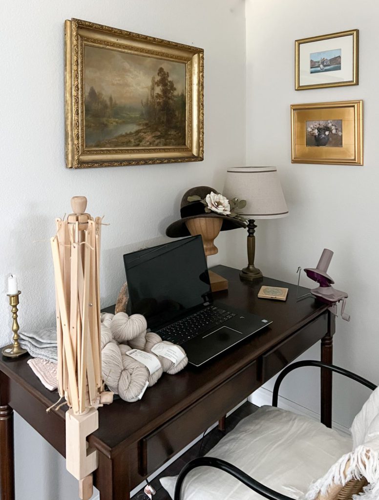 A dark brown desk with lots of knitting design tools (including a swift, ball winder, and skeins of yarn) sits tucked into a corner with three pieces of art on the wall.