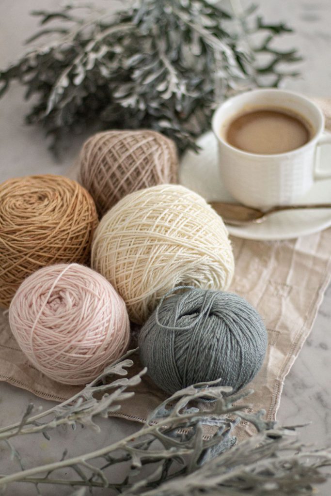 A sideways photo of five balls of yarn in light blue, pale pink, white, a warm tan, and a cool tan. They are surrounded by dusty miller and a white teacup filled with coffee. The balls of yarn are in the middle of the photo, with dusty miller in the foreground and background, and the teacup slightly blurred in the background.