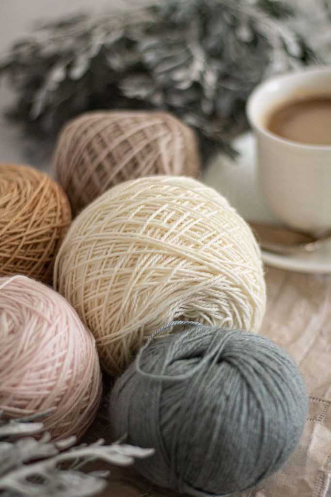 A zoomed-in, sideways photo of five balls of yarn in light blue, pale pink, white, a warm tan, and a cool tan. They are surrounded by dusty miller and a white teacup filled with coffee.