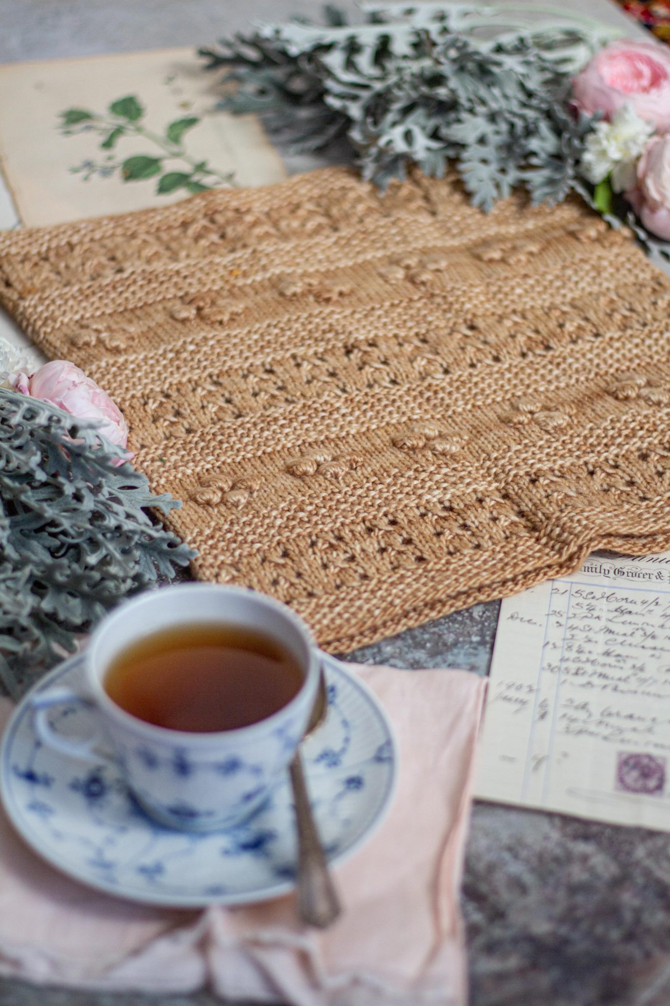 A golden-colored cowl with alternating rows of eyelets, bobbles, and seed stitch is laid flat and surrounded by pink and white roses, antique paper ephemera, and a blue and white teacup. The photo is shot from a low angle at the bottom left of the cowl, so that the teacup is blurred in the foreground and the cowl is clear in the middle of the photo