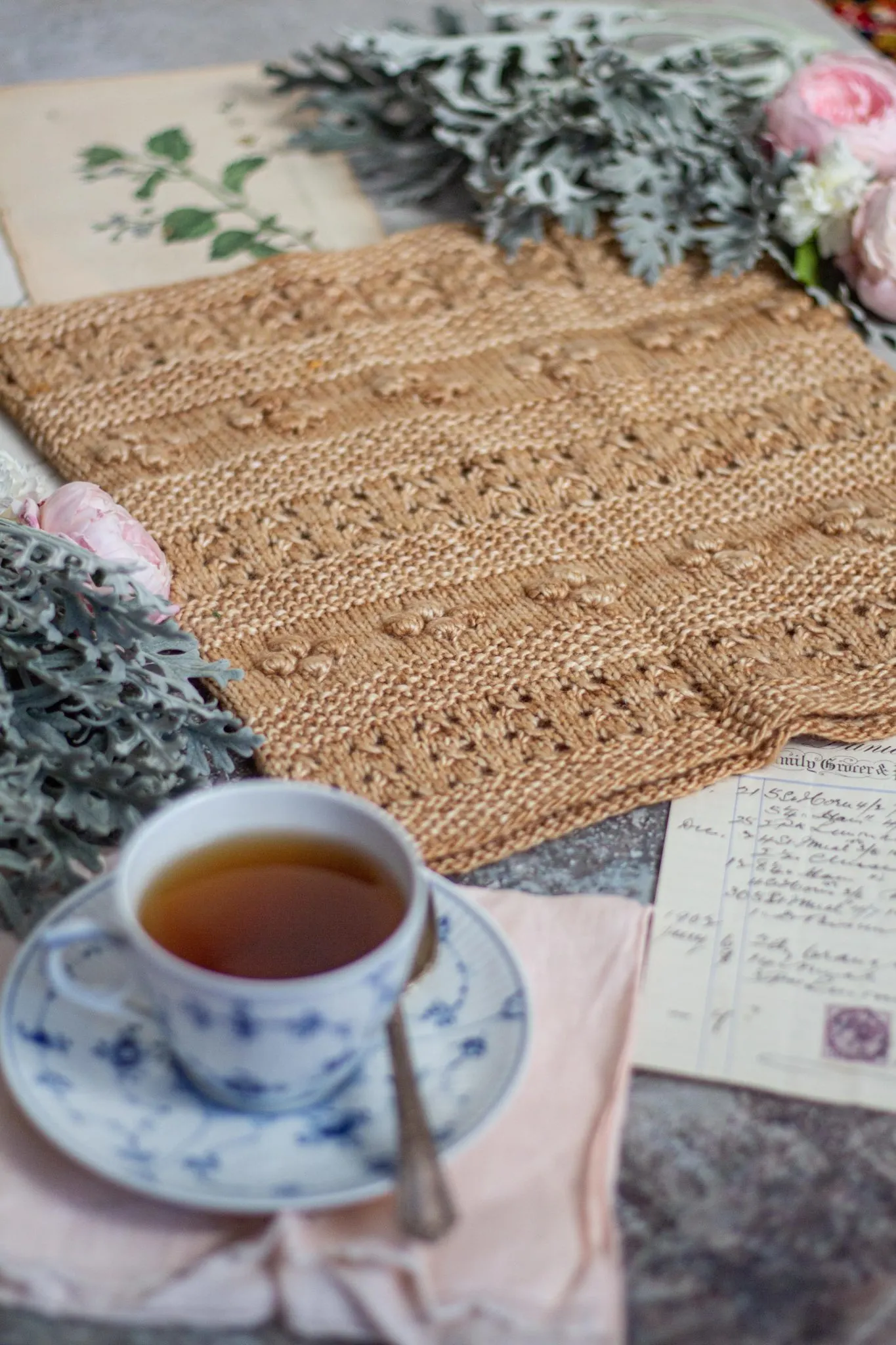 A golden-colored cowl with alternating rows of eyelets, bobbles, and seed stitch is laid flat and surrounded by pink and white roses, antique paper ephemera, and a blue and white teacup. The photo is shot from a low angle at the bottom left of the cowl, so that the teacup is blurred in the foreground and the cowl is clear in the middle of the photo