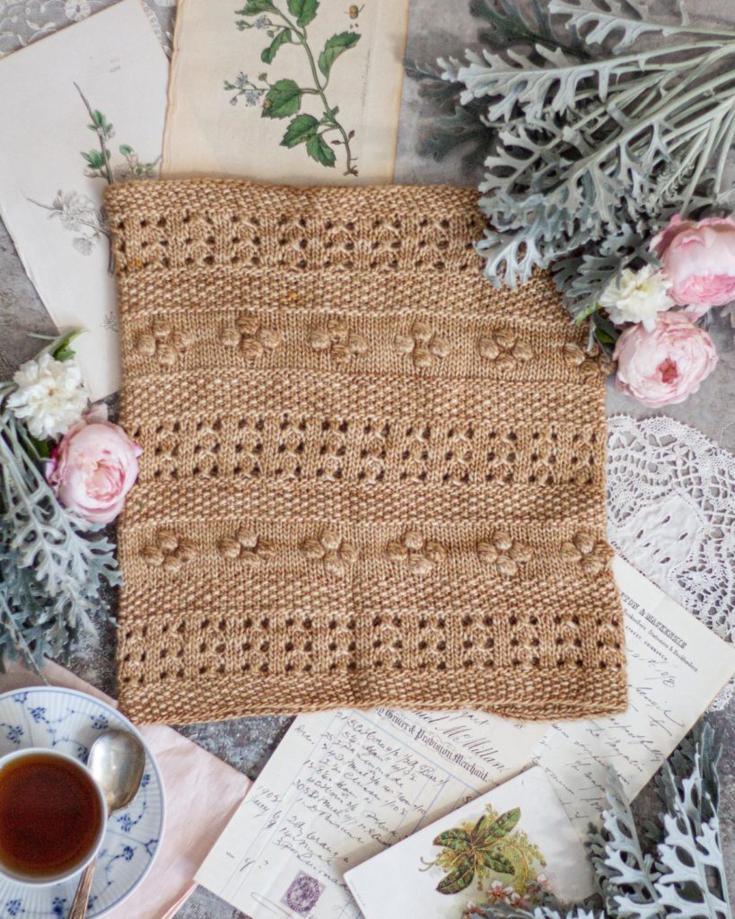 A golden-colored cowl with alternating rows of eyelets, bobbles, and seed stitch is laid flat and surrounded by pink and white roses, antique paper ephemera, and a blue and white teacup.