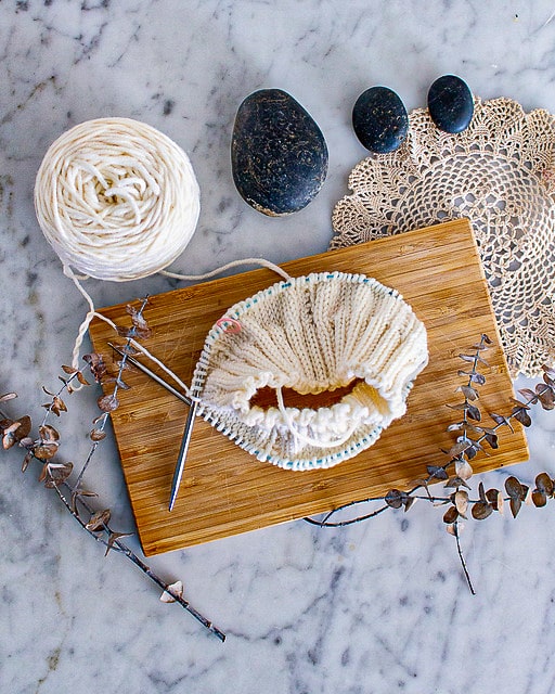 A partially knit white hat on steel circular needles is laid flat on a wooden cutting board surrounded by dried eucalyptus, three dark gray stones, and a cream-colored doily.