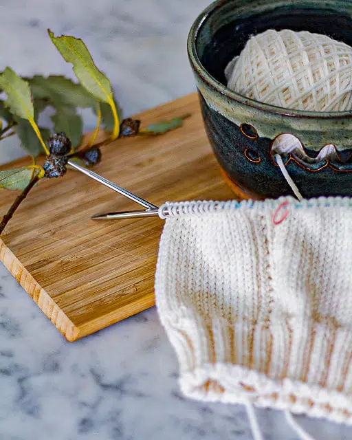 A partially knit white hat on steel circular needles is laid flat on a wooden cutting board. The hat is in the foreground. In the background is a ceramic yarn bowl full of more white yarn and a dried eucalyptus branch.