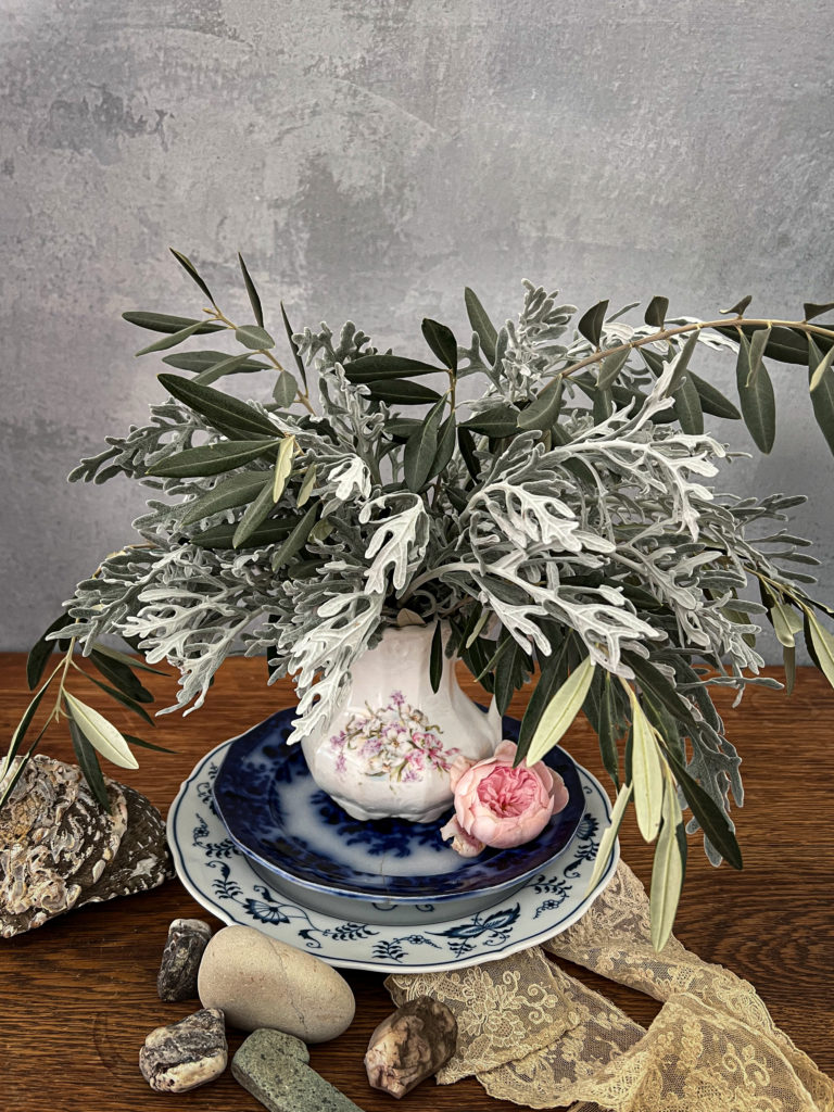 An antique stoneware jug is filled with dusty miller and olive branches sits on top of a small stack of blue and white plates.