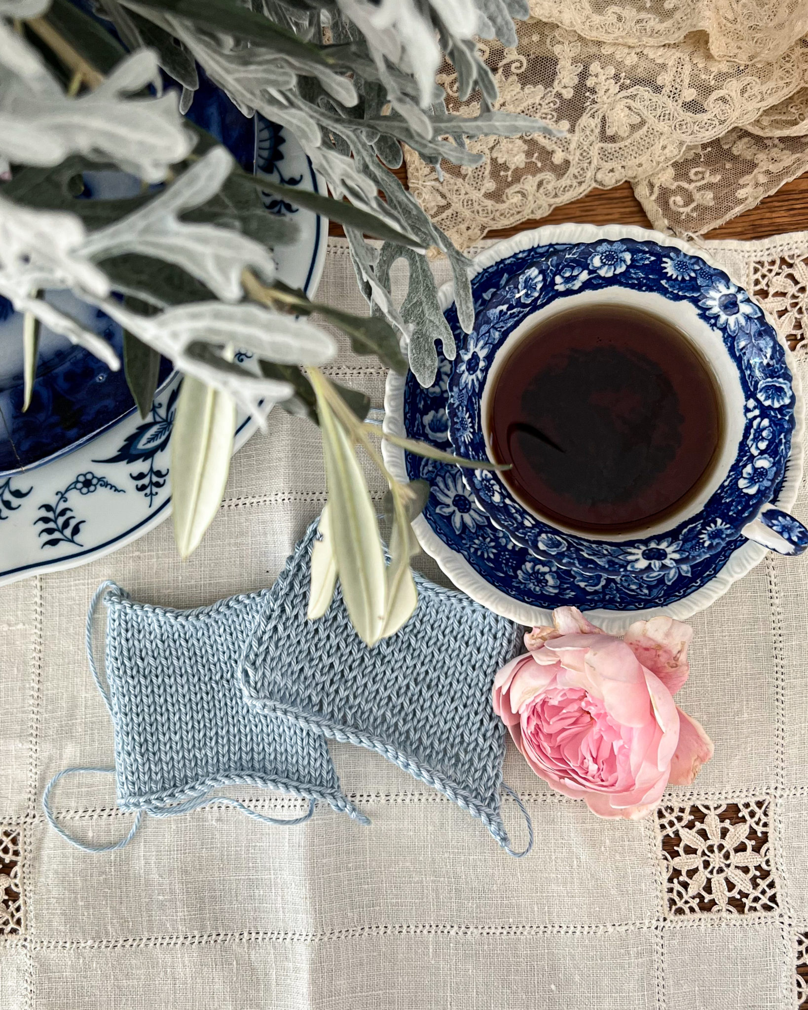 Two blue rectangle swatches are viewed top down through blurred plant leaves in the foreground. One swatch has even stitches, while one has visible rowing out. They are on a white linen placemat next to a blue and white teacup.