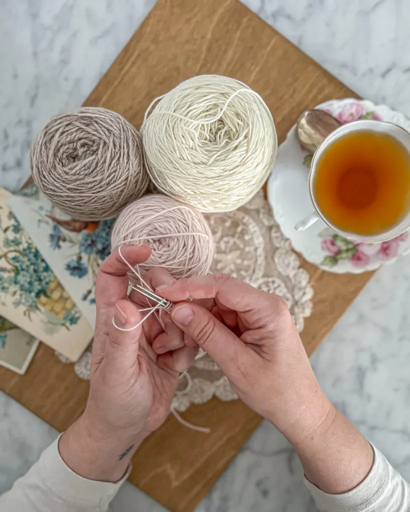 A top-down view of two small, plump, white hands casting on yarn over two needles. Also in the photo is a wooden board, some balls of pastel yarn, and a pink and white teacup.
