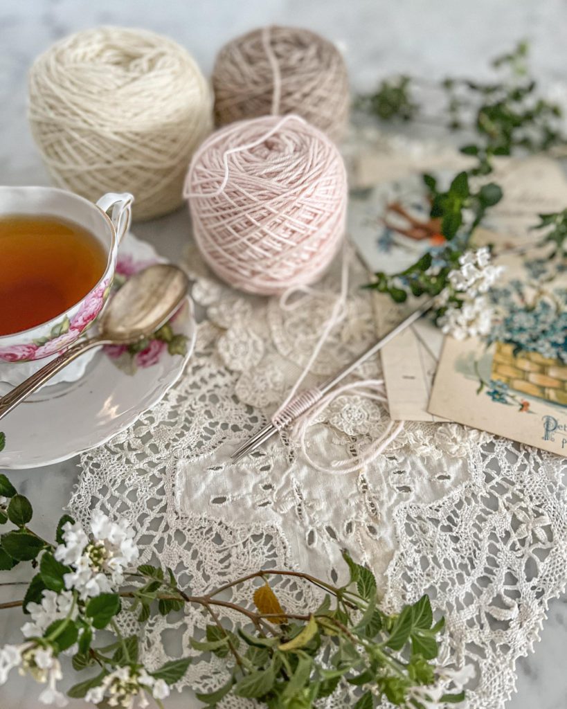A sidelong view of pale pink yarn cast onto two small needles. Also in the photo are some sprigs of greenery, other balls of cream and tan yarn, and a pink and white teacup and saucer.