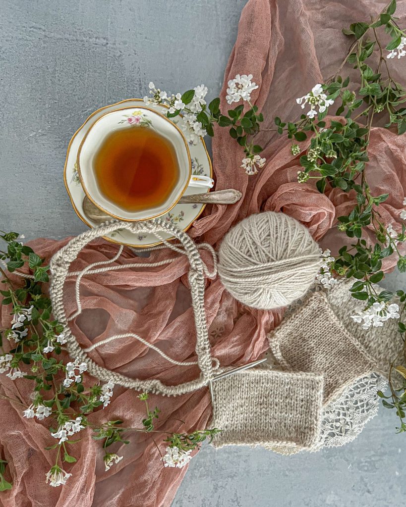 A top-down photo of cream-colored yarn in a ball with some swatches and a new project just cast on to circular needles. The yarn is all laid out on top of a deep pink gauze fabric on a gray background. To the top left are a teacup and saucer, and there are several sprigs of greenery at the edges.