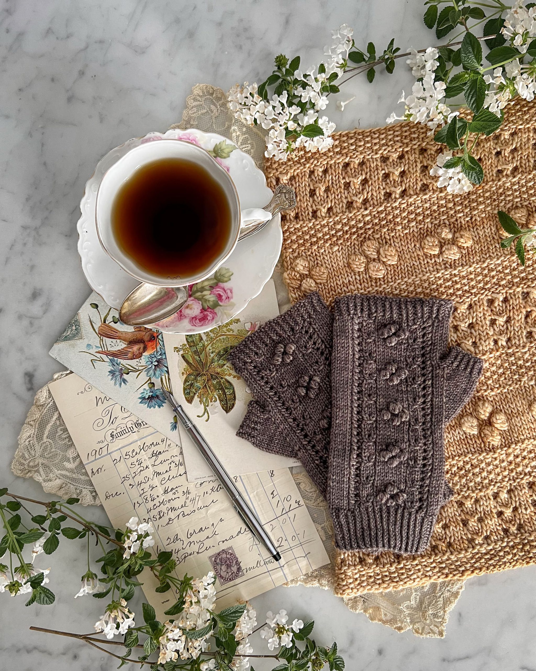 A top-down shot of a pair of brown fingerless mitts resting on top of a golden cowl, both adorned with many bobbles. To the side are antique paper ephemera and a pink and white teacup.