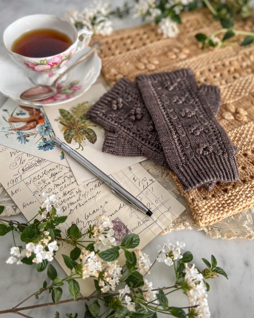 A sideways shot of a pair of brown fingerless mitts resting on top of a golden cowl, both adorned with many bobbles. To the side are antique paper ephemera and a pink and white teacup.