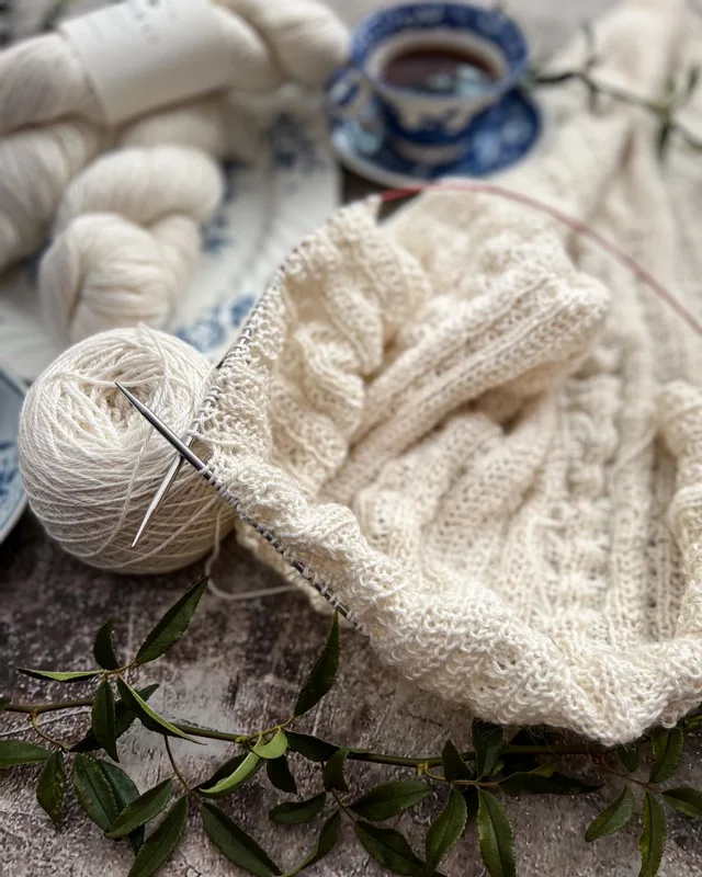 A sideways view of a white lacy wrap, half finished and still on the needles. Blurred in the background is a blue and white teacup and more skeins of white yarn.