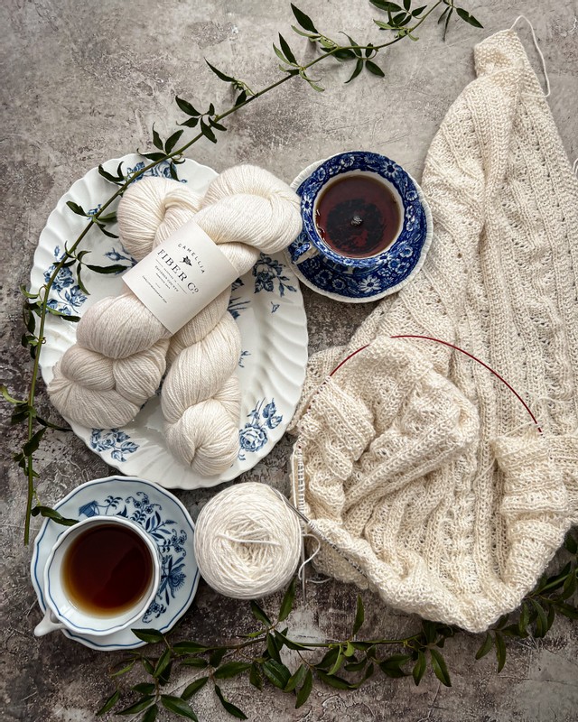 A top-down view of a lacy white wrap, half-knitted and still on the needles. To its left are two blue and white teacups filled with tea, along with two more skeins of white yarn on a blue and white platter.