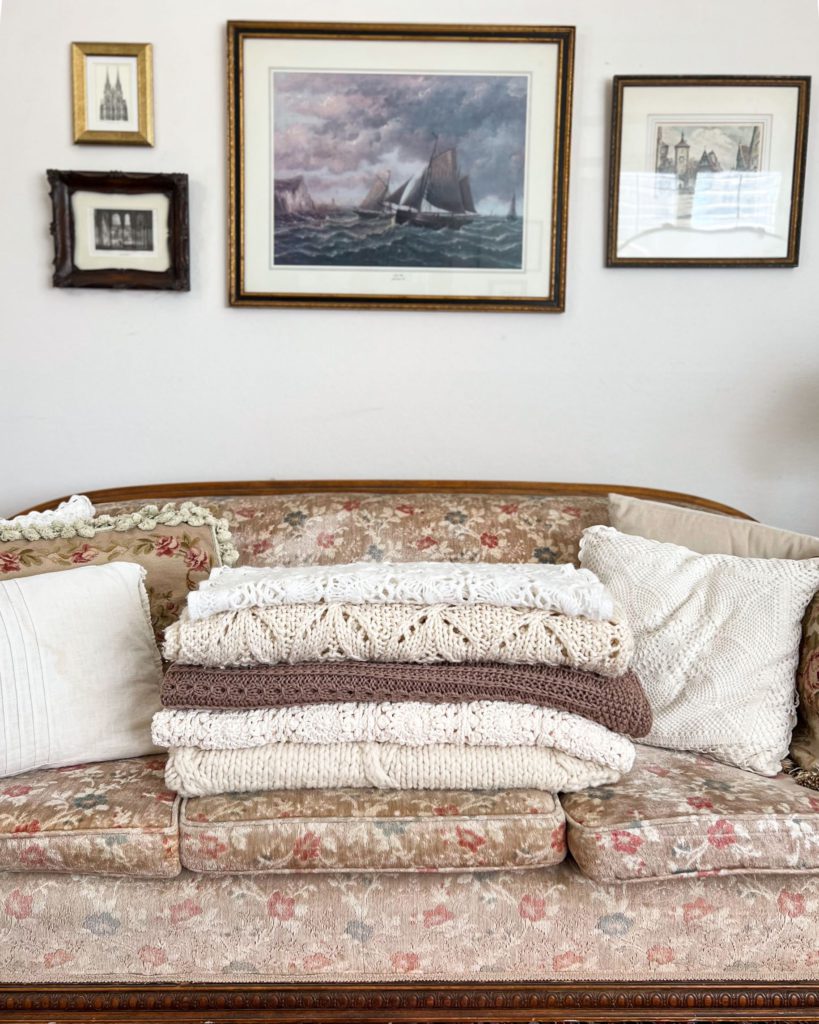 A stack of knit and crocheted blankets in neutral colors on an antique floral sofa
