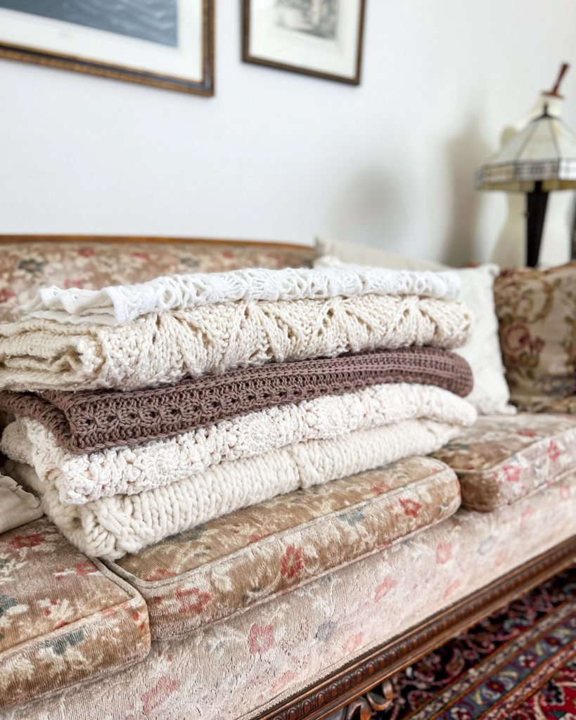 A sidelong image of a stack of neutral-colored knit and crochet blankets on a vintage floral couch.