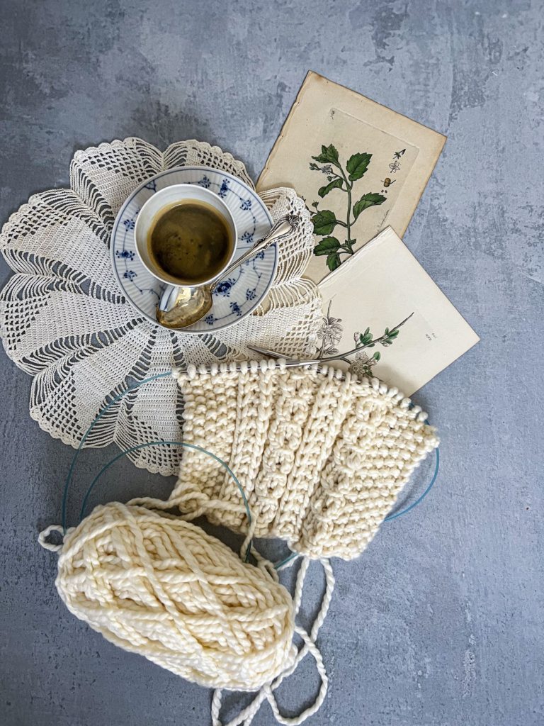 A top-down shot of a cream-colored gauge swatch in progress, along with a teacup, a doily, and some antique botanical prints.