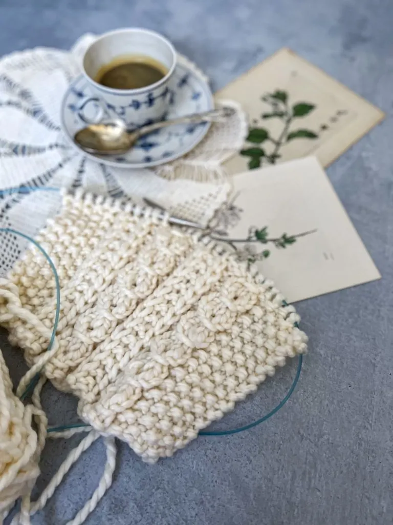 A close-up image of a cream-colored swatch of knit fabric. Behind it, blurred in the background, are a teacup, a doily, and some antique botanical prints.