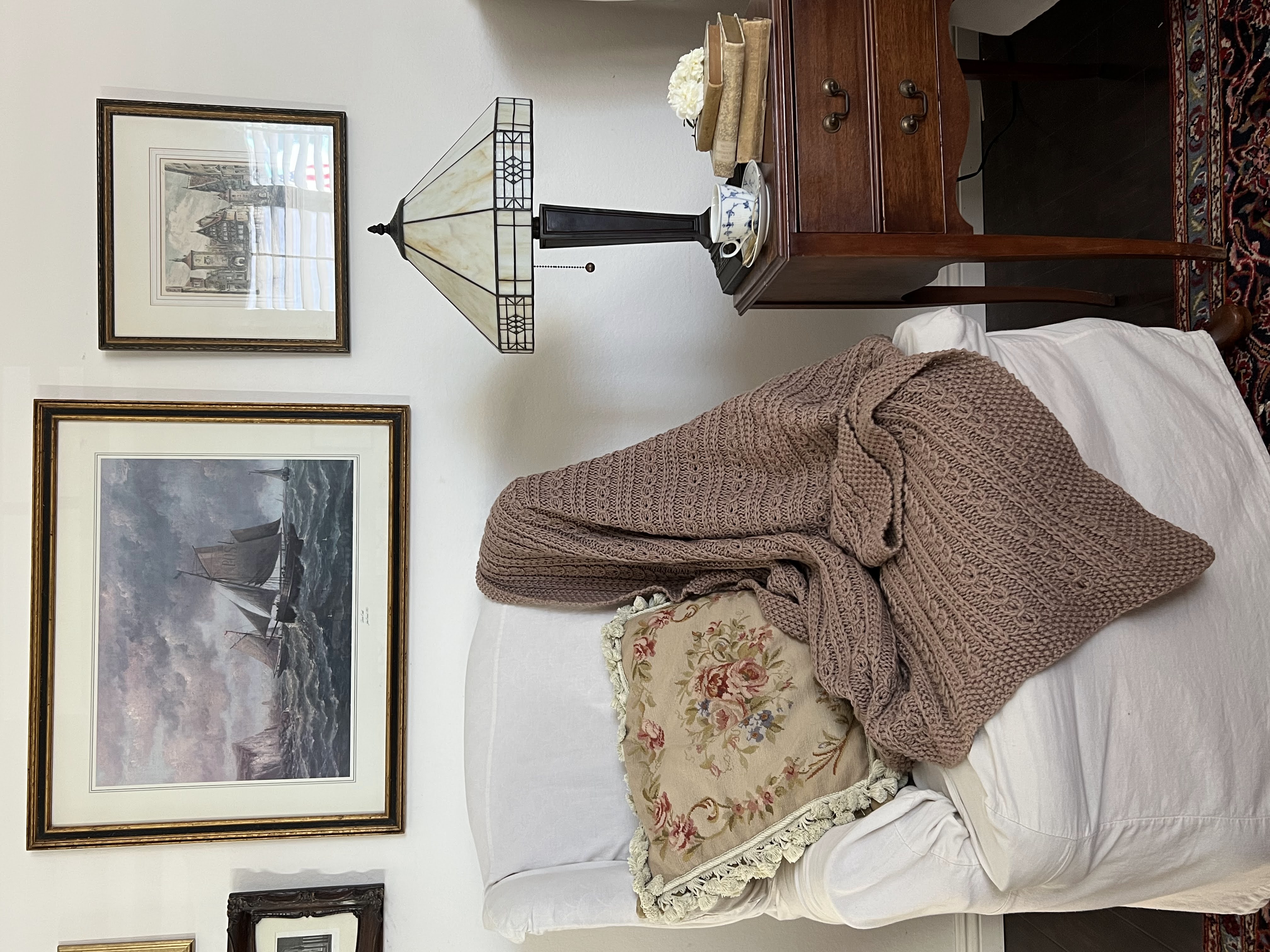 A zoomed out image of the Catalina Eddy Blanket draped across the side and seat of a wing-back chair. Also in the image are an antique table, a glass-shaded lamp, and antique prints on the wall.