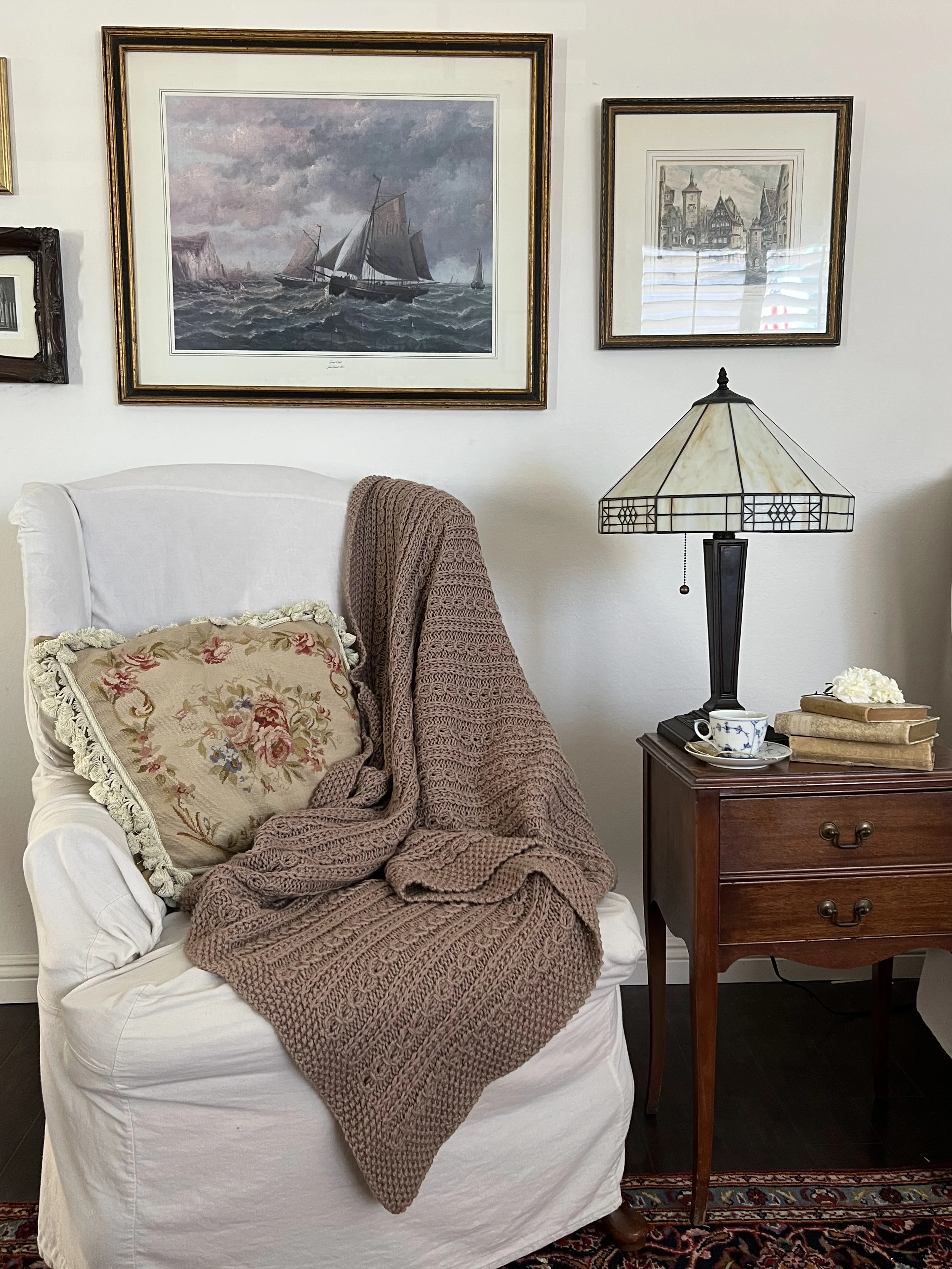 A zoomed out image of the Catalina Eddy Blanket draped across the side and seat of a wing-back chair. Also in the image are an antique table, a glass-shaded lamp, and antique prints on the wall.
