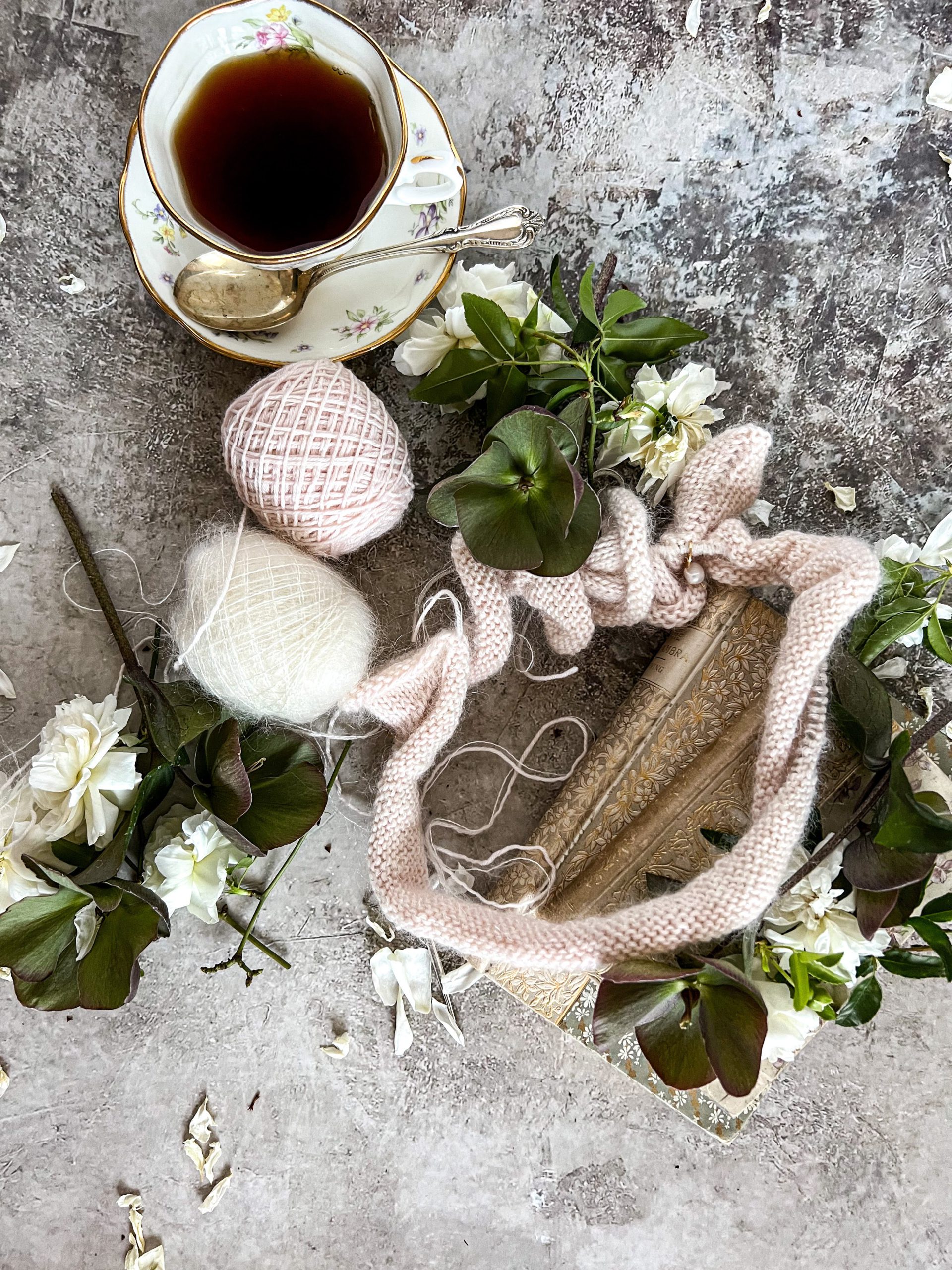 A top-down image of pink and white balls of yarn, some pink knitting on a circular needle, a cup of tea, and some antique books surrounded by flowers