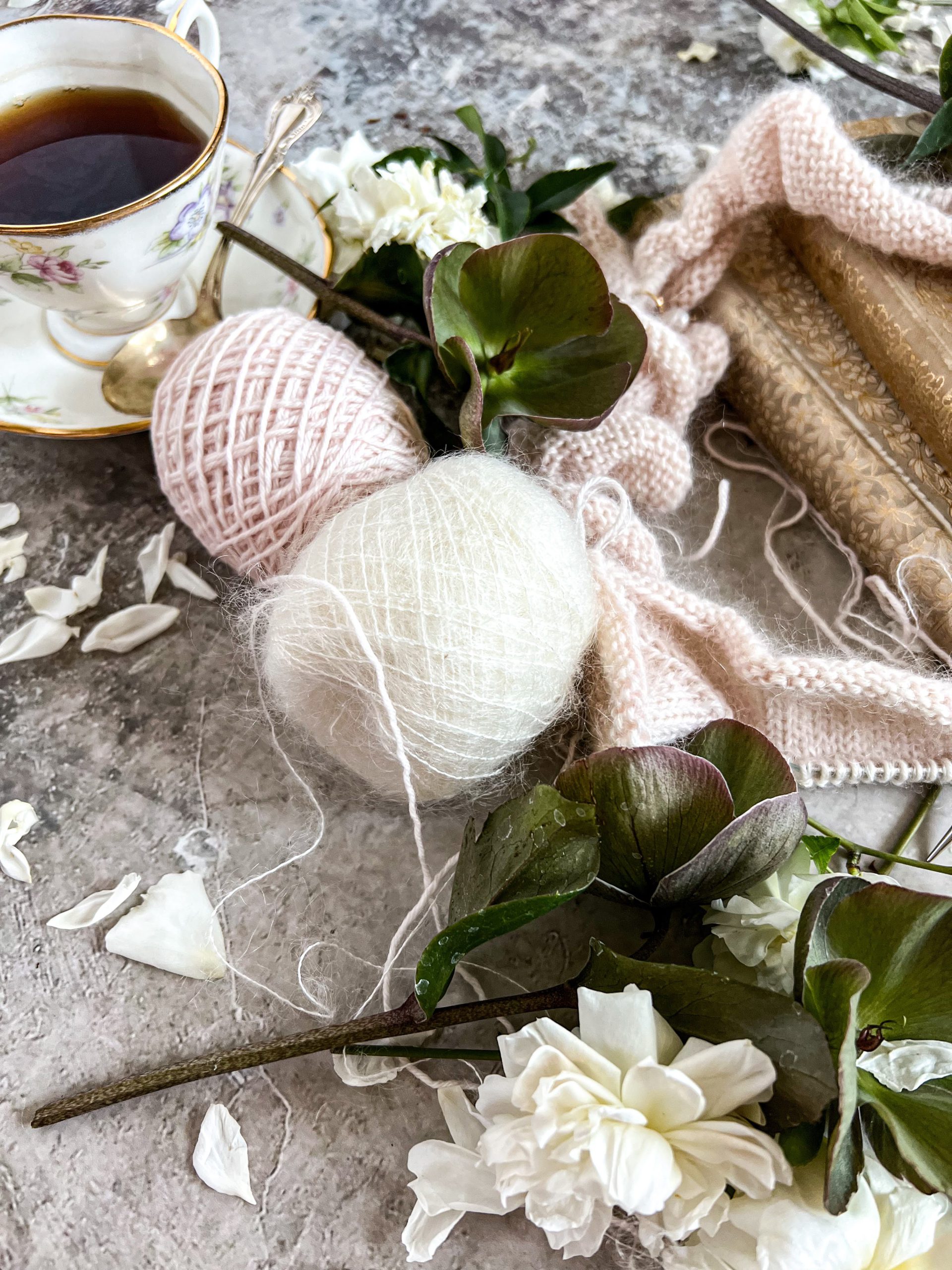A close-up on two balls of yarn, one pink and one white, surrounded by flowers, a teacup, and some knitting