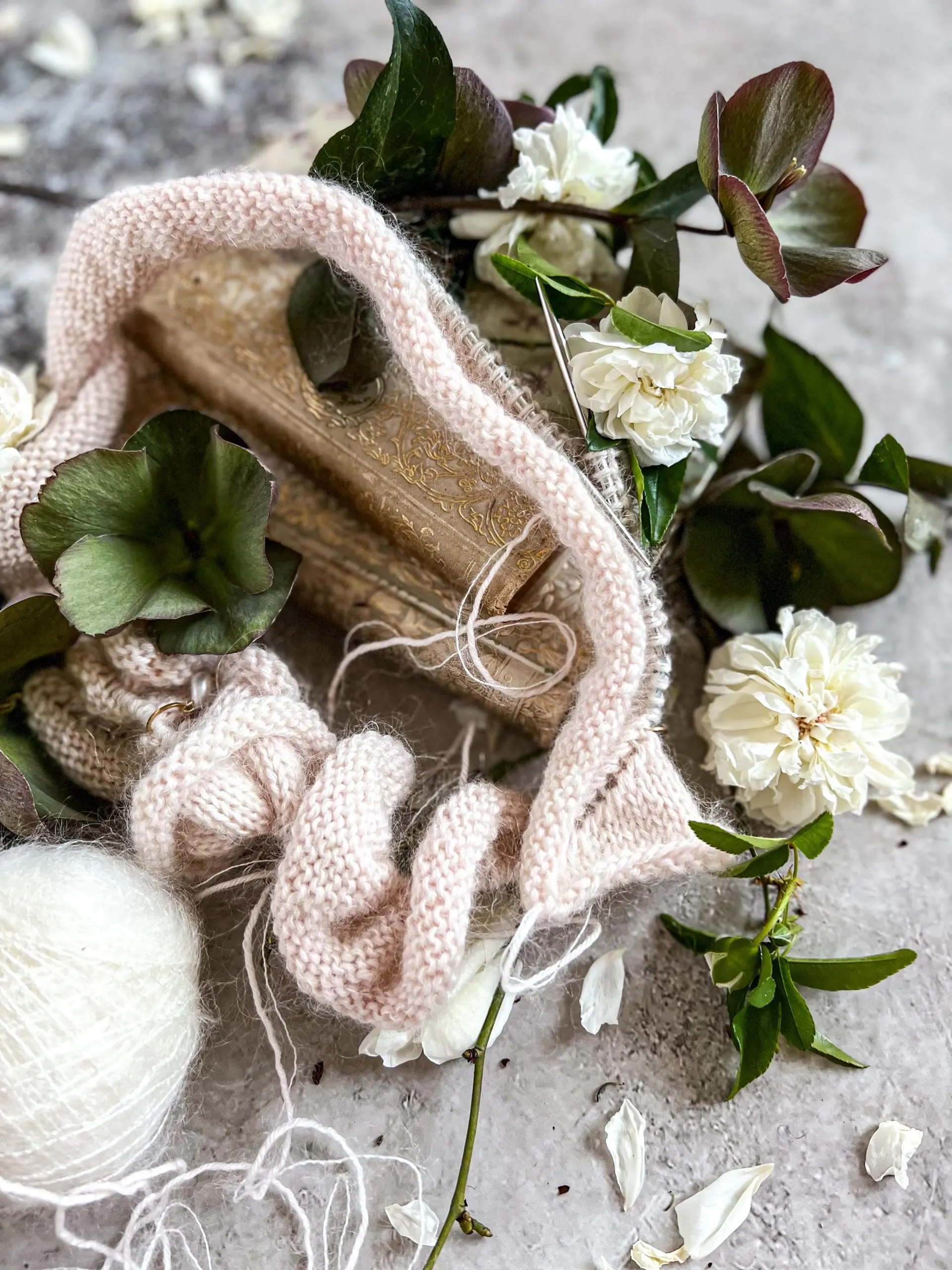 Pink knitting on a circular needle surrounded by hellebores, white rosettes, and antique books