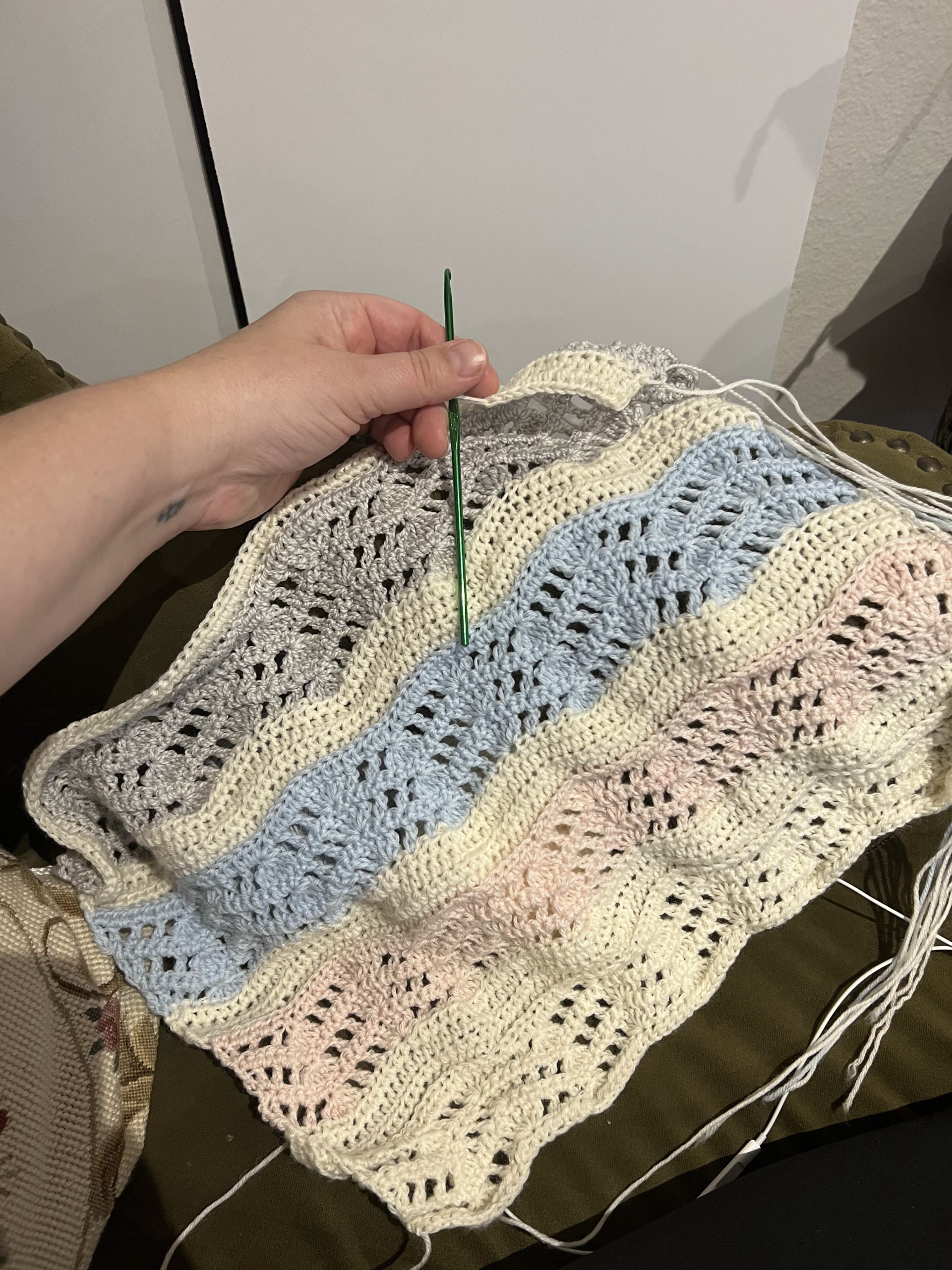A partially finished lacy crochet wrap with stripes in cream, pink, blue, and gray. My left hand with the crochet hook is visible at top left.