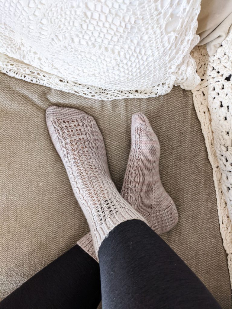 The Lucida Socks, a pair of lavender-colored socks with panels of eyelets and cables running down front and back, are modeled on a pair of feet crossed at the ankles on a sofa