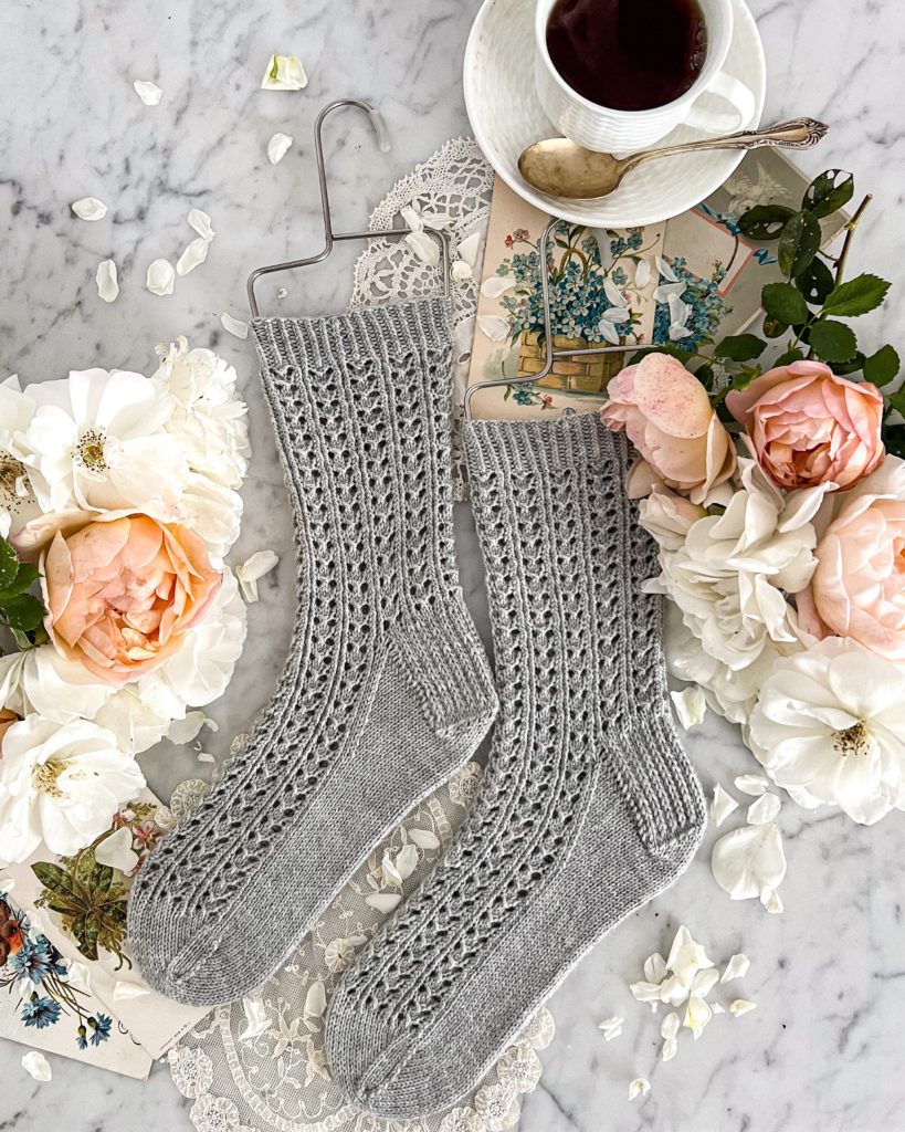 A top-down view of a pair of gray, lacy socks laid out flat on a white marble counter. They're surrounded by roses, antique paper ephemera, and a white teacup full of coffee.