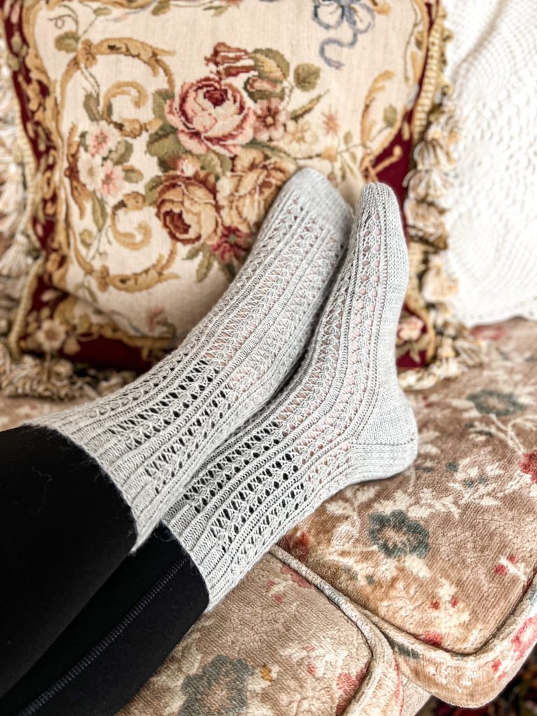 A pair of gray, lacy socks on two feet that are crossed at the ankles, photographed from the side.