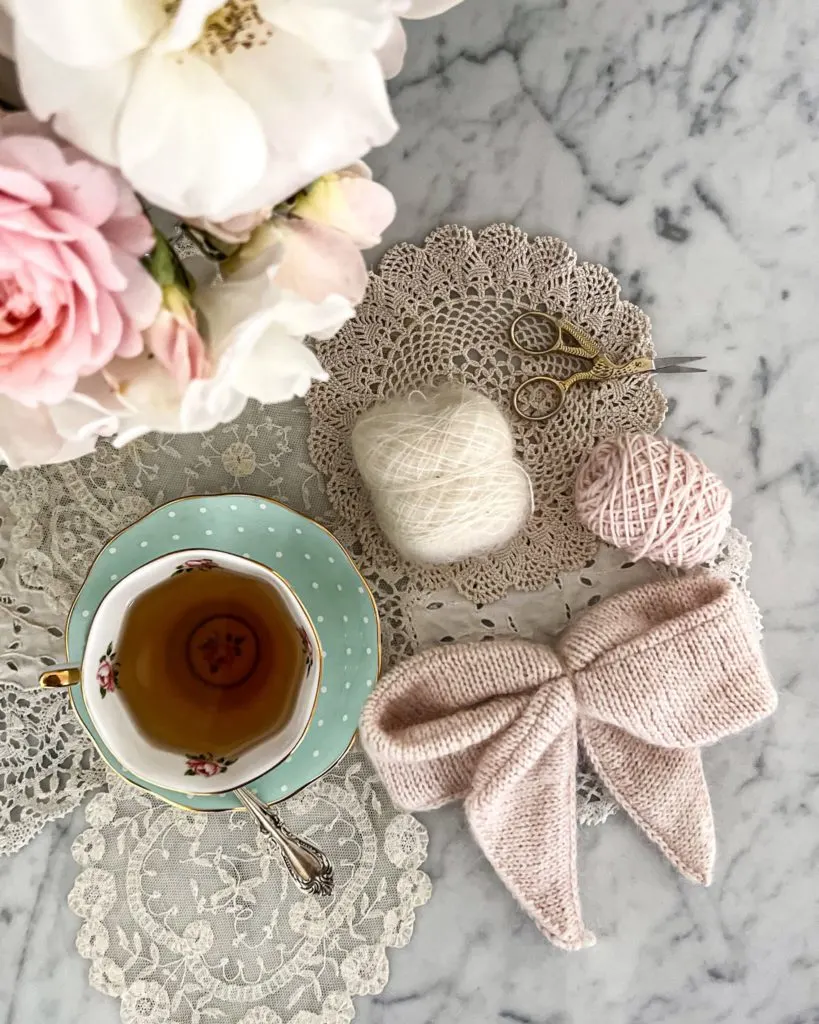 A top-down image of a pink, fuzzy, knit bow, surrounded by some small balls of yarn and a teacup full of tea