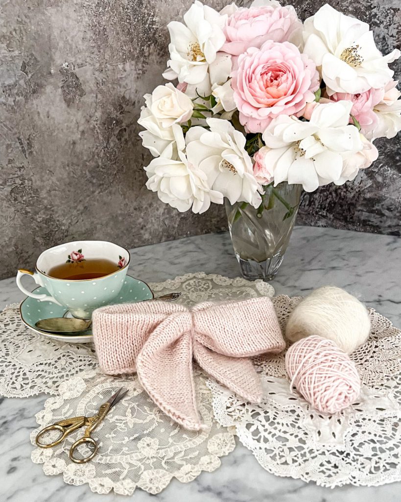 A pink, fuzzy, knit bow sits in the center of the photograph, surrounded by a vase of pink and white roses, some small balls of yarn, and a polka dotted teacup