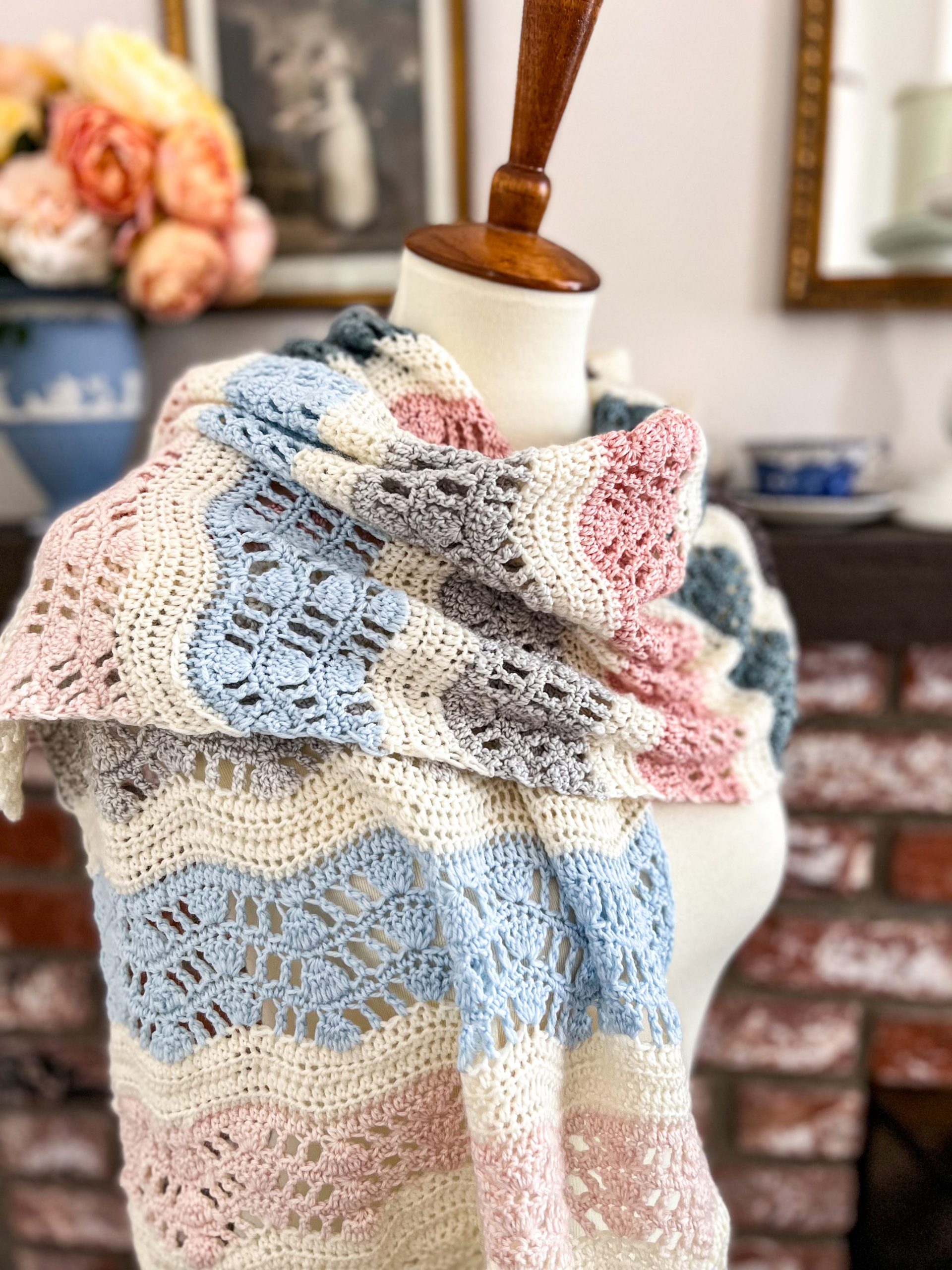 A close-up on the Intentions Wrap, showing off its lacy openwork and stripes in pale pink, white, blue, and gray. The wrap is draped around the shoulders of a white dressmaker's form.