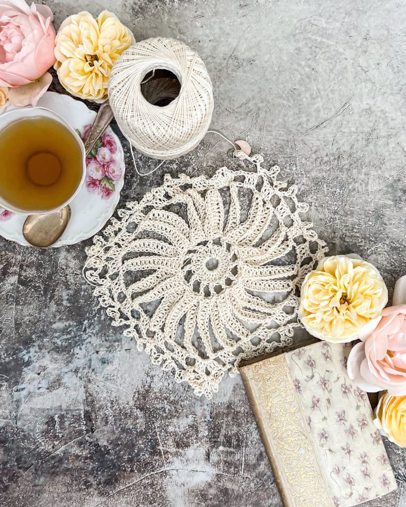 A top-down image of a loosely crocheted round starburst shape. Arranged with it are some pink and yellow roses, an antique book, and a teacup full of chamomile.