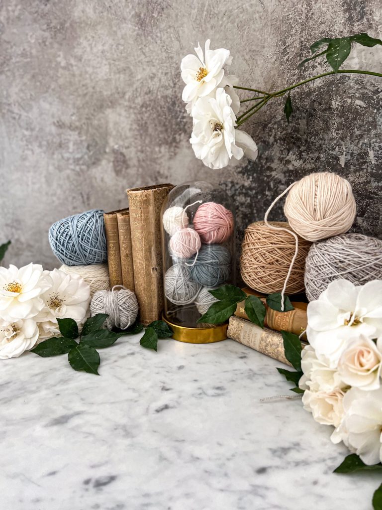 A side-angle shot of a collection of yarn, antique books, and white roses arranged in a gentle curve against a gray background. In the center is a glass cloche full of small yarn balls.